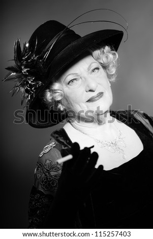 Good looking senior woman glamour vintage style. Holding a cigarette. Wearing a black hat. Holding a black fan. Black and white studio shot. Short blonde curly hair. Chic look. Dressed in black.