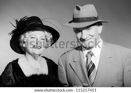 Good looking senior couple glamour vintage style. Wearing a hat. Black and white studio shot. Short blonde curly hair. Chic look. Dressed in black. Wearing a grey suit with tie.