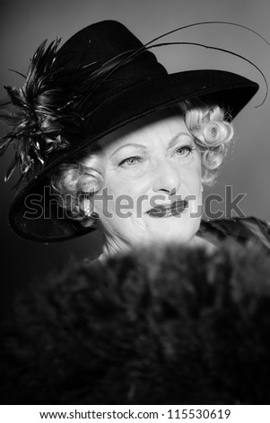 Good looking senior woman glamour vintage style. Wearing a black hat. Holding a black fan. Black and white studio shot. Short blonde curly hair. Chic look. Dressed in black.