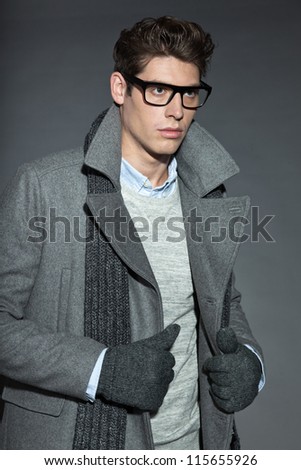 Men winter fashion. Handsome man with brown hair wearing grey scarf, grey sweater, grey coat, black glasses and grey woolen gloves. Casual look. Studio shot isolated on grey background.