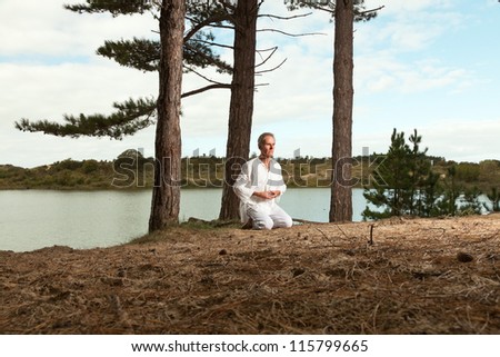 Senior spiritual man dressed in white. Short grey hair. Meditating in nature. Outdoors. Forest. Dunes. Cloudy sky. Healthy living.