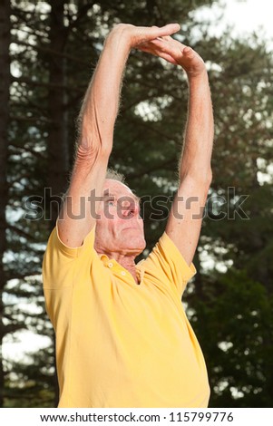 Senior man stretching his arms. Doing workout in nature. Healthy living. Forest. Outdoors. Yellow shirt and white pants. Short grey hair.