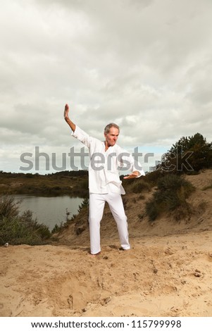 Senior spiritual man dressed in white. Short grey hair. Working out in nature. Outdoors. Dune landscape. Cloudy sky. Healthy living.