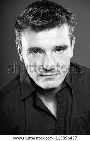 Mean looking handsome man with short brown hair wearing black shirt. Good looking. Fashion studio portrait. Isolated on grey background. Glamour black and white portrait.
