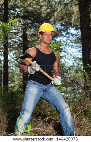 Muscled lumberjack with black shirt and axe in forest. Wearing yellow safety helmet.