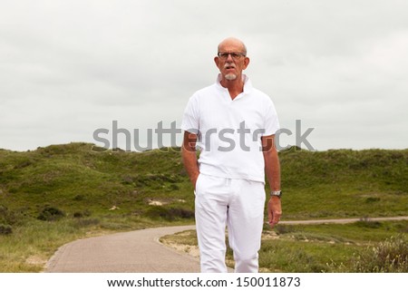 Retired senior man with beard and glasses walking outdoors in grass dune landscape with cloudy sky.