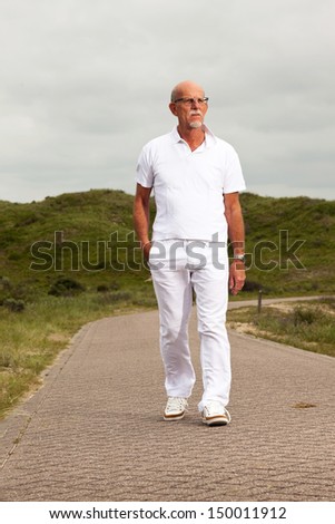 Retired senior man with beard and glasses walking outdoors in grass dune landscape with cloudy sky.