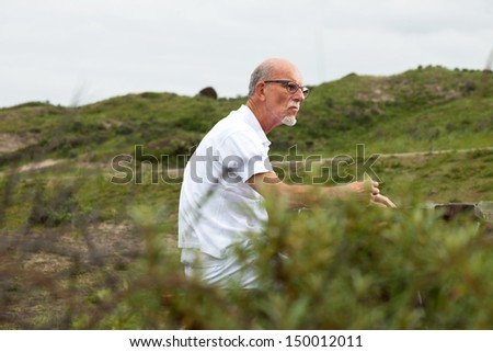 Retired man with beard and glasses resting and having lunch in grass dune landscape. Dressed in white.