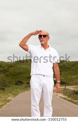 Retired senior man with beard and sunglasses walking outdoors in grass dune landscape with cloudy sky.