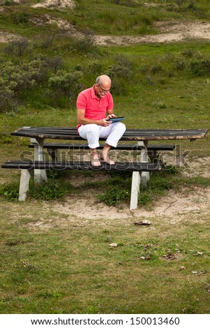 Retired senior man resting and using his tablet at table in park outdoors in grass dune landscape.