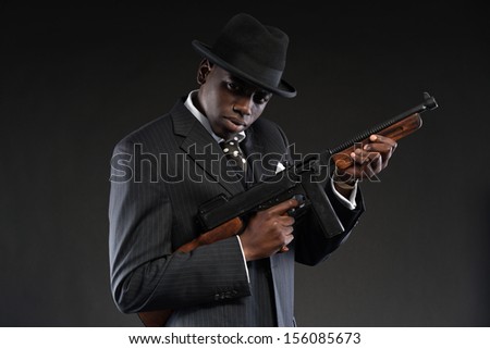 Retro african american mafia man wearing striped suit and tie and black hat. Holding machine gun. Studio shot.