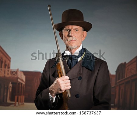Senior western man wearing a brown hat and coat holding rifle. Standing in old small cowboy town.