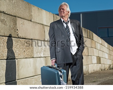 Depressed senior business man without a job and homeless on the street. Holding a suitcase. Wearing a dirty suit.