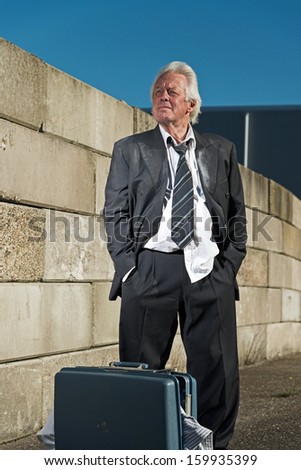 Depressed senior business man with suitcase without a job and homeless on the street. Wearing a dirty suit.