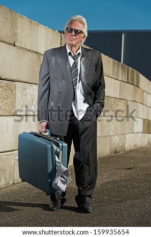 Depressed senior business man with sunglasses without a job and homeless on the street. Holding a suitcase. Wearing a dirty suit.