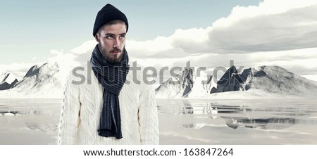 Cool man with beard in winter fashion. Wearing white woolen sweater black cap and grey scarf. Outdoor in snow mountain landscape.