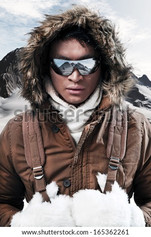 Asian winter sport fashion man with sunglasses and backpack in arctic mountain landscape. Wearing brown jacket with fur hoody and white gloves.