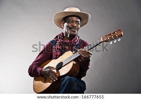 Retro senior afro american blues man in times of slavery. Wearing denim bib and brace overall with straw hat. Playing acoustic guitar.