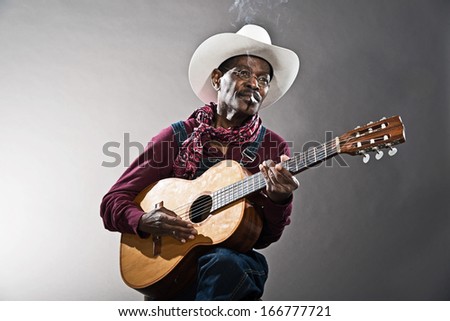 Retro senior afro american blues man in times of slavery. Wearing denim bib and brace overall with white hat. Playing acoustic guitar.