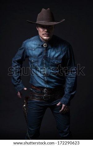 Modern fashion cowboy. Wearing brown hat and blue jeans shirt. Pulling his gun. Blonde hair and beard. Studio shot against black.