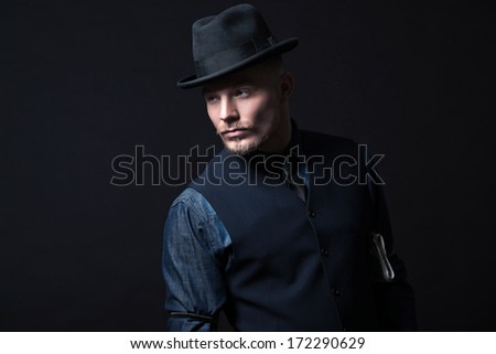 Retro 1900 modern fashion man. Wearing blue jeans shirt with gilet and trousers. Black hat. Studio shot against black.