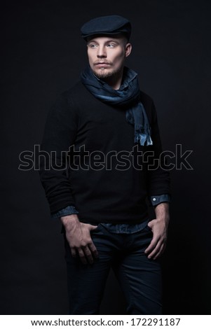 Man winter fashion. Wearing black sweater with scarf and cap. Blonde hair and beard. Studio shot against dark wall.