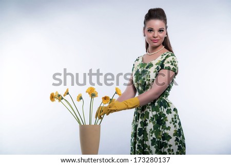Retro 50s fashion housewife wearing yellow rubber gloves. Putting flowers in vase. Studio shot against grey.