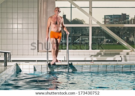 Healthy active senior man with beard in indoor swimming pool going in the water. Wearing orange swimming trunks.