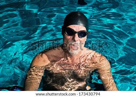 Active healthy senior man with beard in swimming pool wearing black cap and glasses.