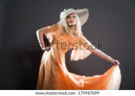 Soft focus retro hippie 70s fashion sensual girl with long blonde hair wearing an orange dress and hat. Studio shot against grey.