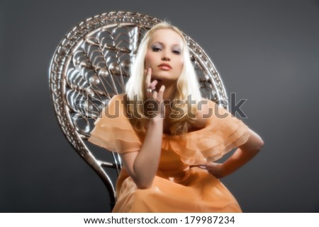 Retro hippie seventies fashion sensual girl with long blonde hair wearing an orange dress. Sitting on a big chair. Studio shot against grey.