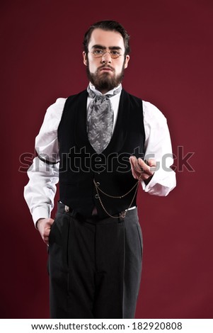 Retro 1900 victorian fashion man with beard wearing black gilet grey tie and glasses. Holding pocket watch. Studio shot against red wall.