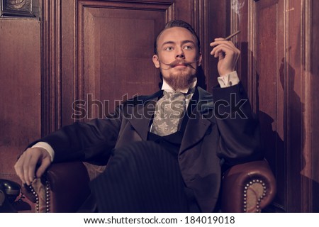 Vintage 1900 fashion man with beard. Sitting in old wooden reading room. Smoking a cigar.
