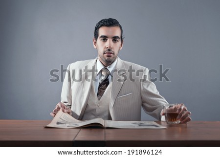 Mafia fashion man wearing white striped suit and tie. Sitting at table with glass of whisky and newspaper. Studio shot.