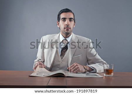 Mafia fashion man wearing white striped suit and tie. Sitting at table with glass of whisky and newspaper. Studio shot.