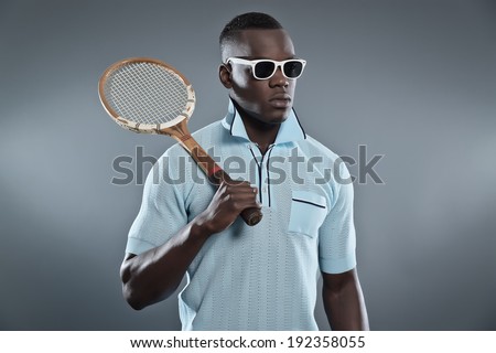 Retro black african tennis player wearing blue shirt and white sunglasses. Holding vintage racket. Studio shot against grey.