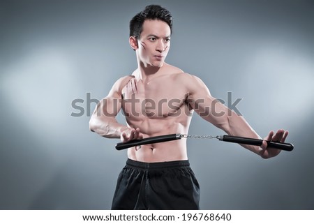 Muscled asian kung fu man in action pose with nunchucks. Blood stripes on his chest and face. Wearing black pants. Studio shot against grey.