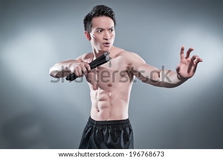 Muscled asian kung fu man in action pose with nunchucks. Blood stripes on his chest and face. Wearing black pants. Studio shot against grey.