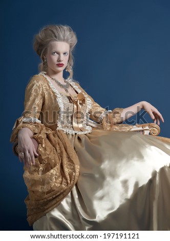 Retro baroque fashion woman wearing gold dress. Sitting on vintage couch. Studio shot against blue.