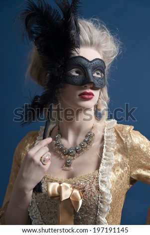 Retro baroque fashion woman wearing gold dress. Holding a black mask. Studio shot against blue.