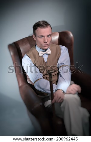 Retro classic business fashion man sitting in big leather chair. Wearing shirt with gilet and bow tie. Holding cane.