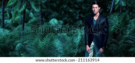 Asian business fashion man in jungle with green ferns and palm trees. Wearing dark grey suit and blue shirt.