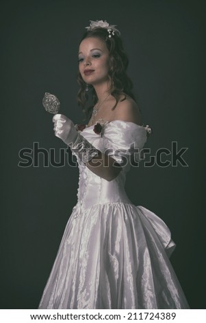 Victorian fashion woman wearing white dress. Holding silver mirror. Studio shot against grey.
