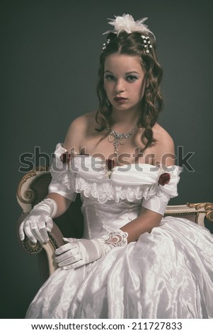Retro victorian fashion woman wearing white dress. Sitting on antique couch. Studio shot against grey.