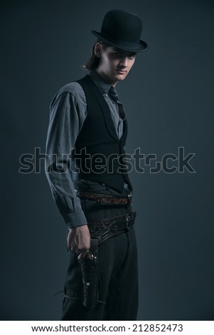 Western 1900 fashion man with brown hair and hat holding gun. Studio shot against grey.