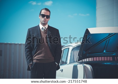 Retro fifties mafia fashion man standing next to open trunk of vintage car.