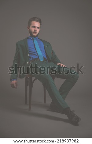 Business fashion man wearing green suit with blue shirt and tie. Sitting on wooden chair. Studio shot against grey.