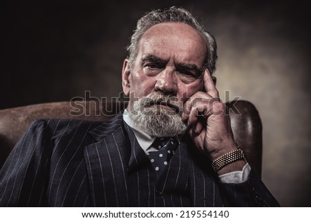 In chair sitting characteristic senior business man. Gray hair and beard wearing blue striped suit and tie. Against brown wall.