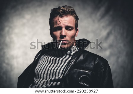 Cigarette smoking retro fifties cool rebellion fashion man wearing striped woolen sweater and black leather jacket. Gray wall.