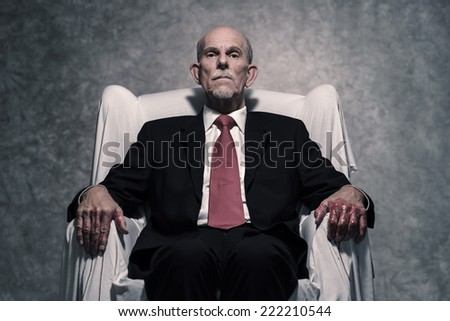 Corrupt businessman with bloody hands sitting in white chair. Gray beard wearing dark suit and red tie. Against grey wall.